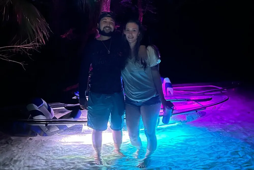 A man and a woman are smiling for a photo at night standing on a beach with colorful lighting and kayaks in the background