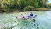 Two people are smiling and kayaking in a clear water body, possibly a river, surrounded by greenery and visible fish beneath them.