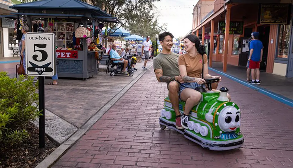 Two people share a light moment as they ride on a whimsical train-shaped scooter down a pedestrian street passing by a speed limit sign indicating 5 mph in an area called Old Town