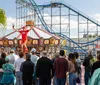 A crowd is watching a performer who is balancing on a unicycle and juggling clubs at an amusement park with a carousel and rollercoaster in the background
