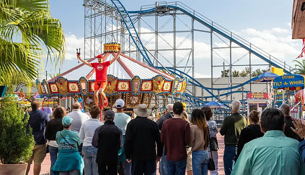 A crowd is watching a performer who is balancing on a unicycle and juggling clubs at an amusement park with a carousel and rollercoaster in the background