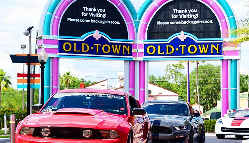 A vibrant archway with the words Old Town stands over a lineup of modern muscle cars under a sunny sky