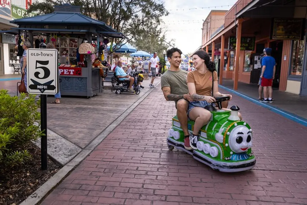 A smiling couple is enjoying a ride on a whimsical green and white train-shaped scooter down a lively street with a 5 MPH speed limit sign on display