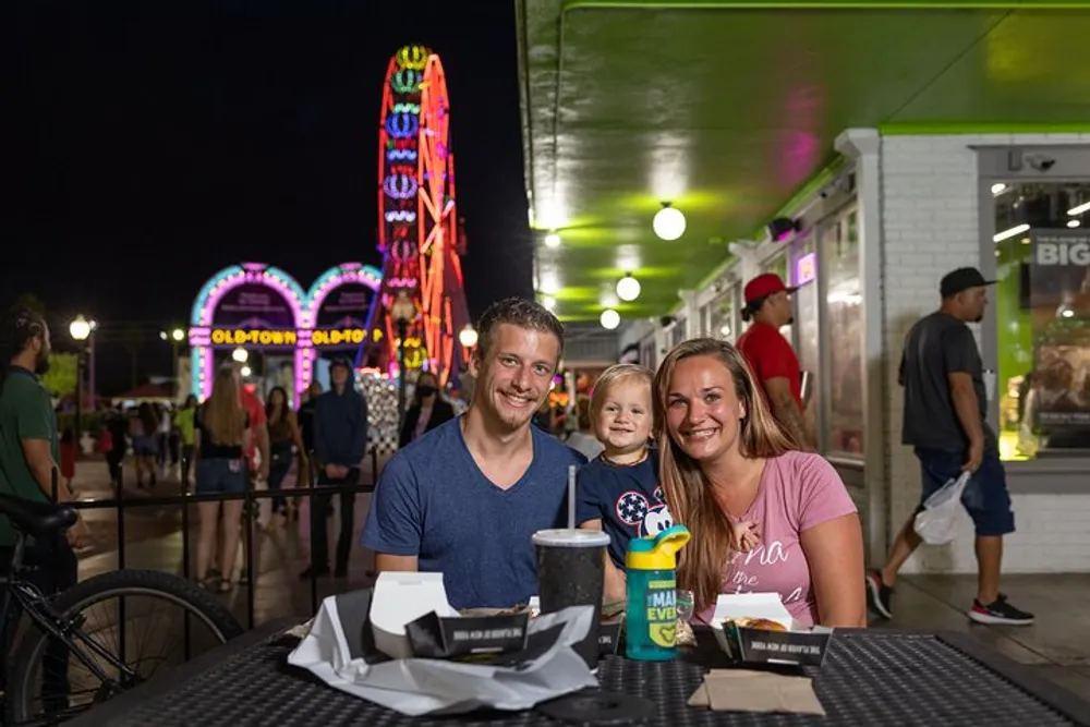 A family with a young child is smiling at the camera while sitting at an outdoor table at a vibrant fairground at night