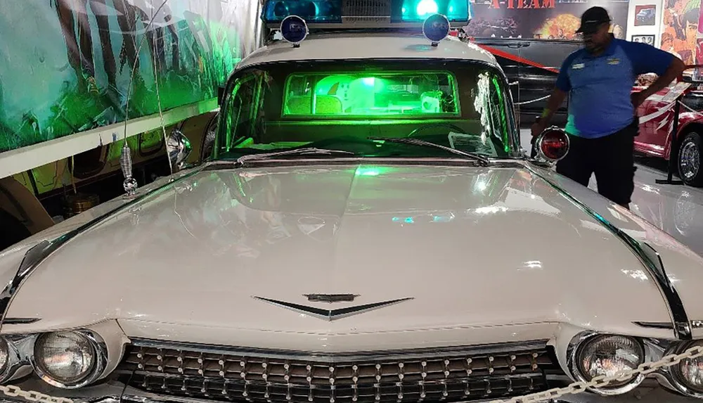 A vintage white police car with a green light inside is displayed in a museum with a person standing beside it against a backdrop of movie memorabilia