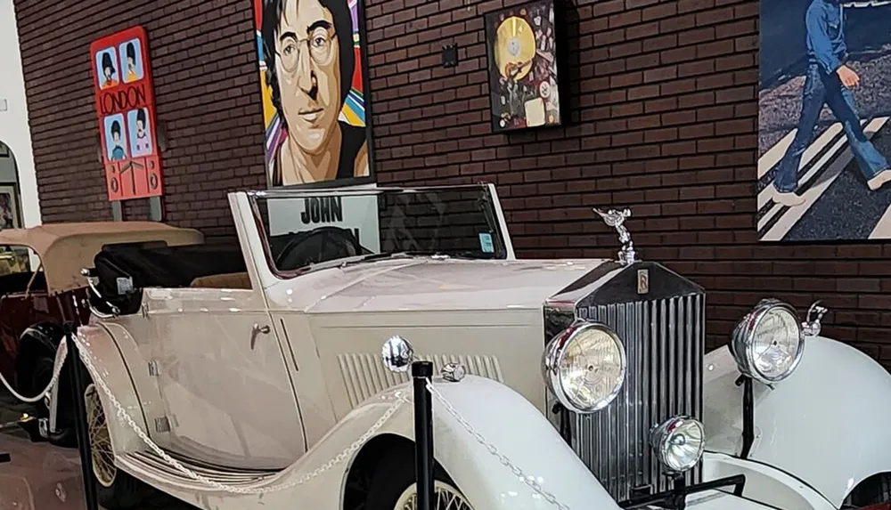 A classic white Rolls-Royce is on display in an indoor setting with a portrait of a man resembling John Lennon on the wall behind it