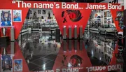 The image shows an exhibition space filled with James Bond memorabilia, including a car, movie posters, and other themed displays, with a large quote saying