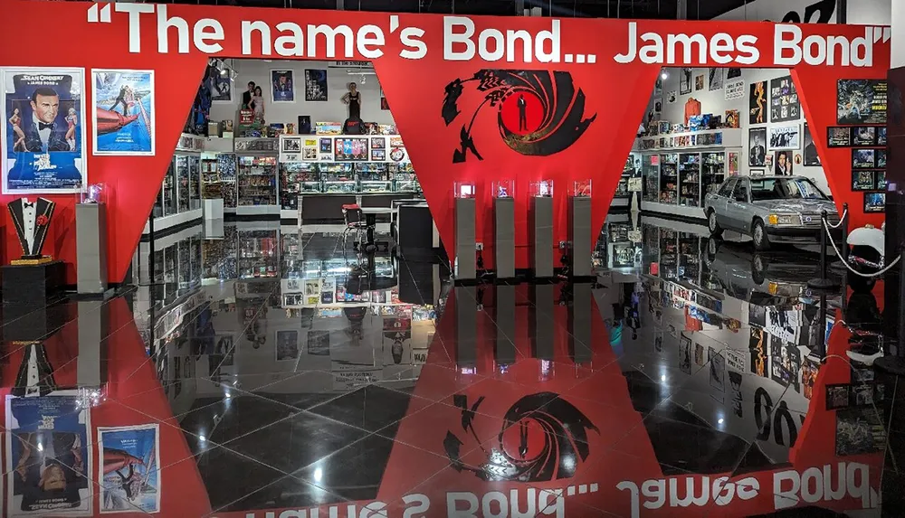 The image shows an exhibition space filled with James Bond memorabilia including a car movie posters and other themed displays with a large quote saying The names Bond James Bond prominently featured