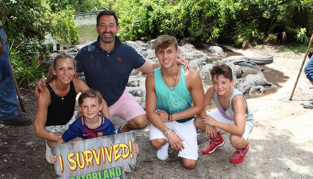 A family is posing with smiles for a photo behind a sign that reads I SURVIVED at a location called Gatorland with several alligators in the background