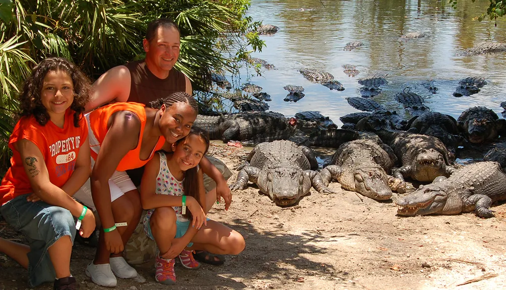 A group of people is posing for a photo near several alligators resting by a body of water