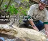 A man wearing a hat and outdoors apparel is crouching next to an albino alligator in a swampy environment smiling at the camera