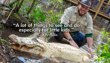 A man wearing a hat and outdoors apparel is crouching next to an albino alligator in a swampy environment, smiling at the camera.