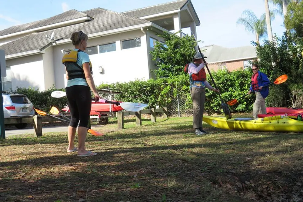 A person wearing a life jacket and cap appears to be instructing or demonstrating something to another person holding a paddle next to a yellow kayak while a spectator watches in a grassy area with kayaks and cars in the background