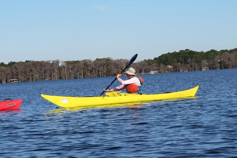 A person is paddling a yellow kayak on calm waters with trees and a clear sky in the background