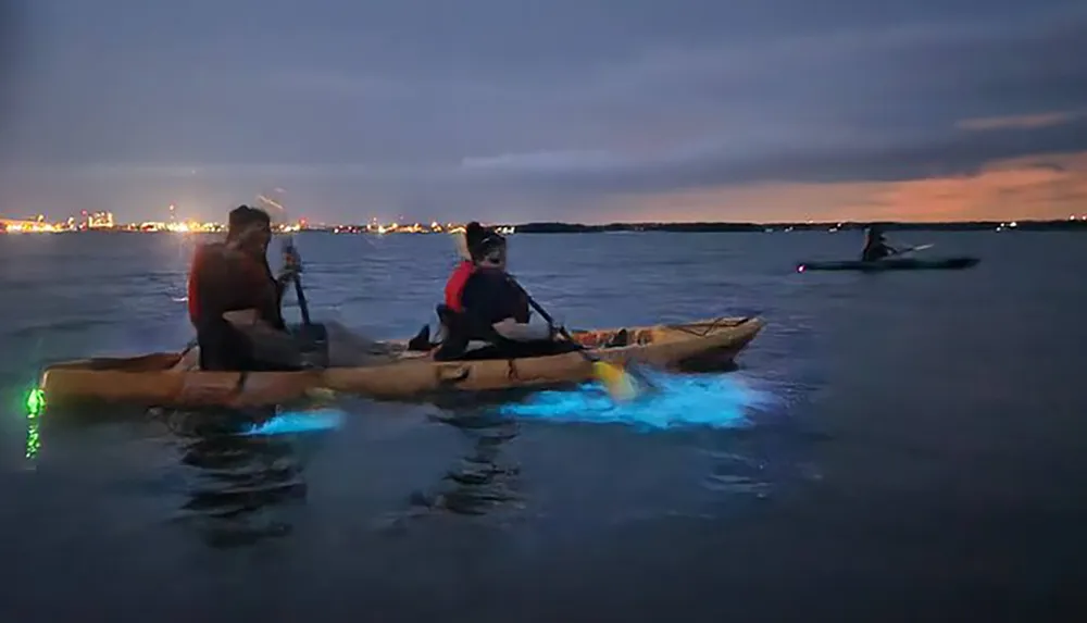 People are kayaking at dusk with illuminated kayaks that cast a blue glow on the water