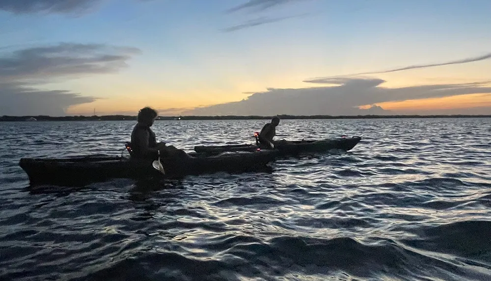 Two people are kayaking on a calm sea as dusk sets in with a colorful twilight sky in the background
