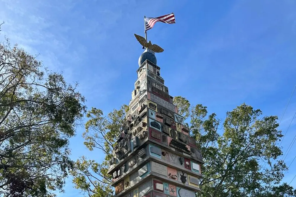 This image features a towering structure adorned with various objects topped by a golden eagle and an American flag fluttering against a blue sky with trees in the background