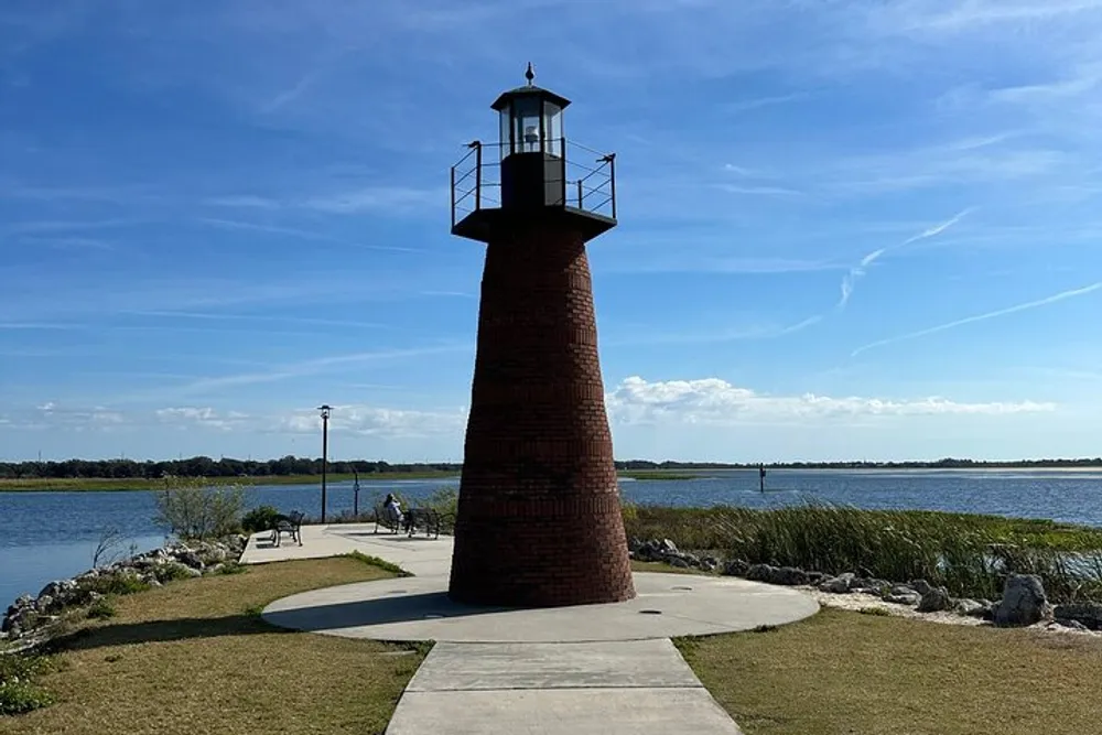 A small brick lighthouse stands by a serene lakeside path under a clear blue sky with people enjoying the pleasant surroundings