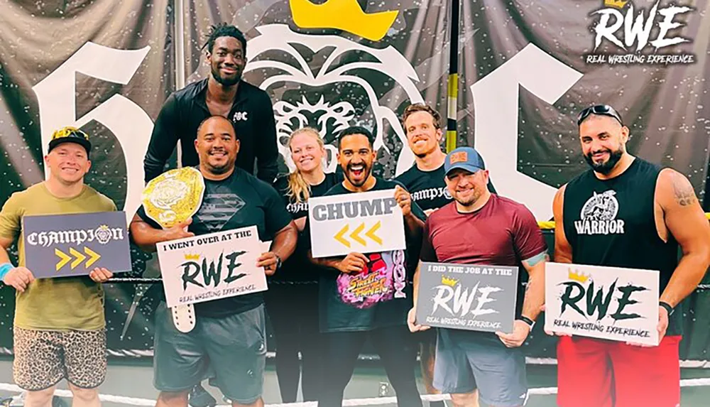 A group of eight smiling people is posing with wrestling-related signs some holding titles in front of a backdrop with a wrestling logo