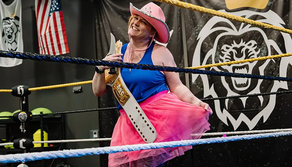 A person in a pink cowboy hat and a tutu smiles while holding a championship belt in a wrestling ring
