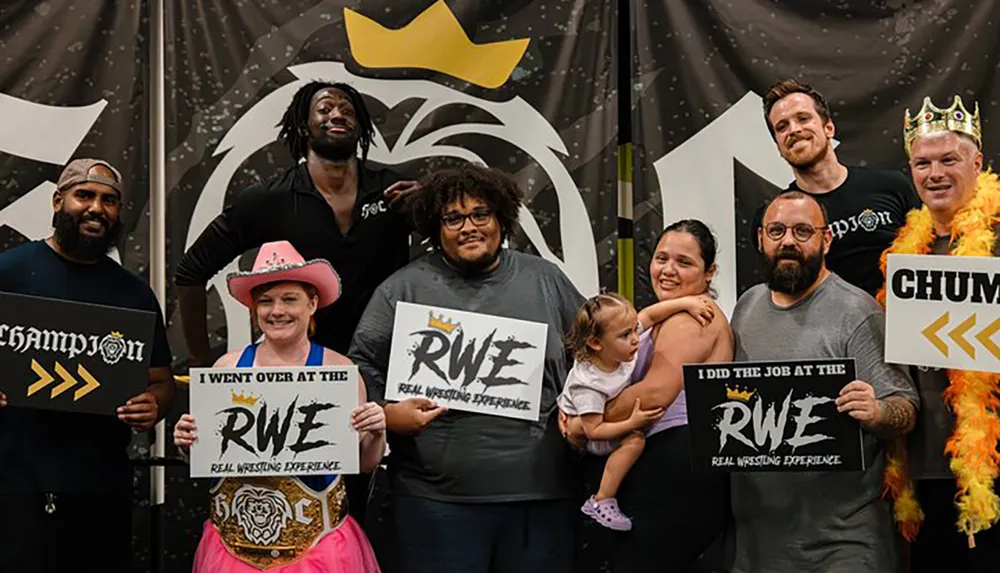 A diverse group of people are posing happily with signs and props related to wrestling in front of a backdrop featuring wrestling-related graphics