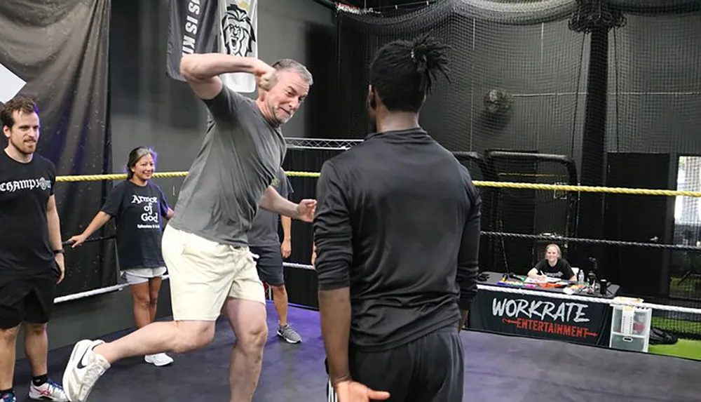 A man is playfully throwing a fake punch at another person inside a training area with onlookers watching and smiling