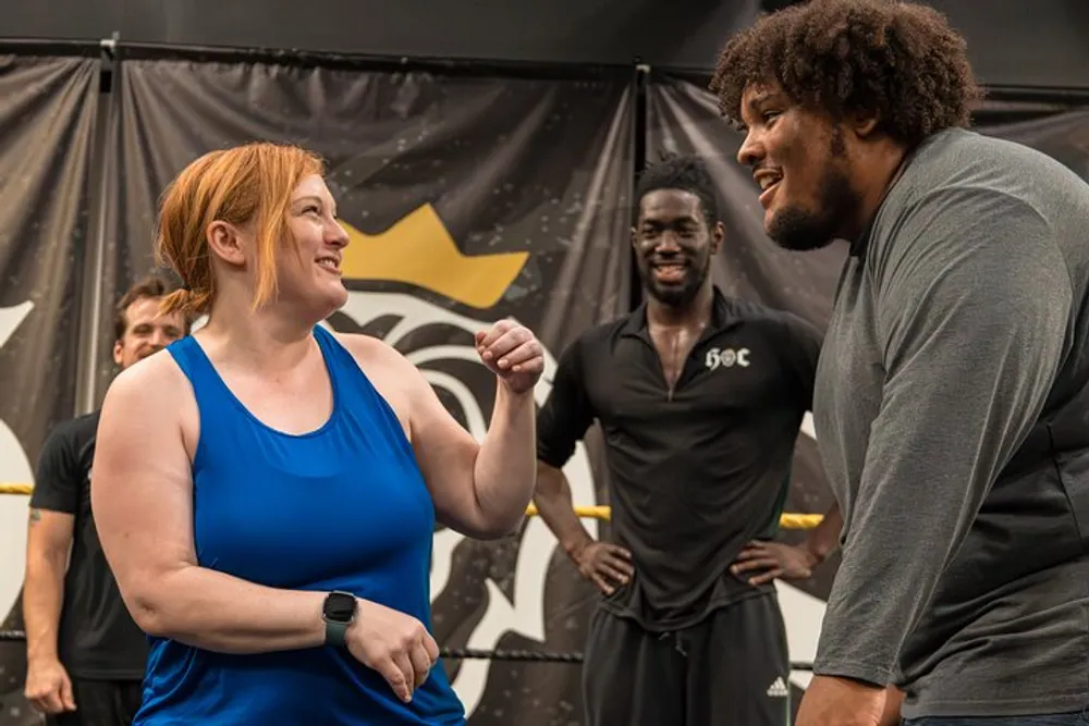 In the image a woman in blue sportswear is facing a man in dark clothing and both are smiling and engaging intently with each other while a second smiling man in the background watches them in what appears to be a gym setting with exercise mats and a logo on the wall