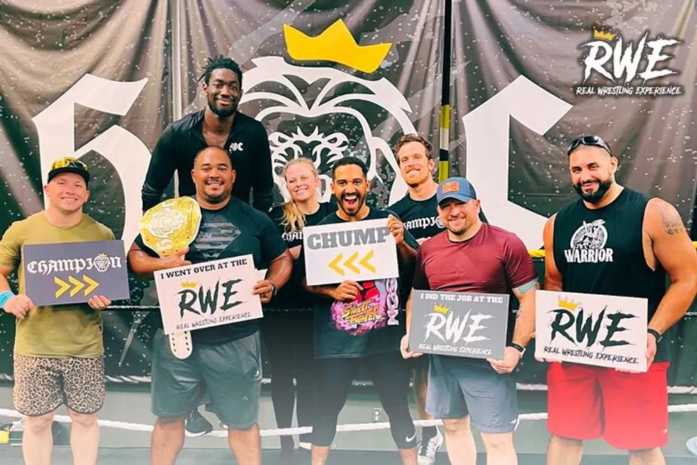 A group of eight smiling people are posing with playful signs and a wrestling belt in front of a backdrop with the logo RWE - Real Wrestling Experience