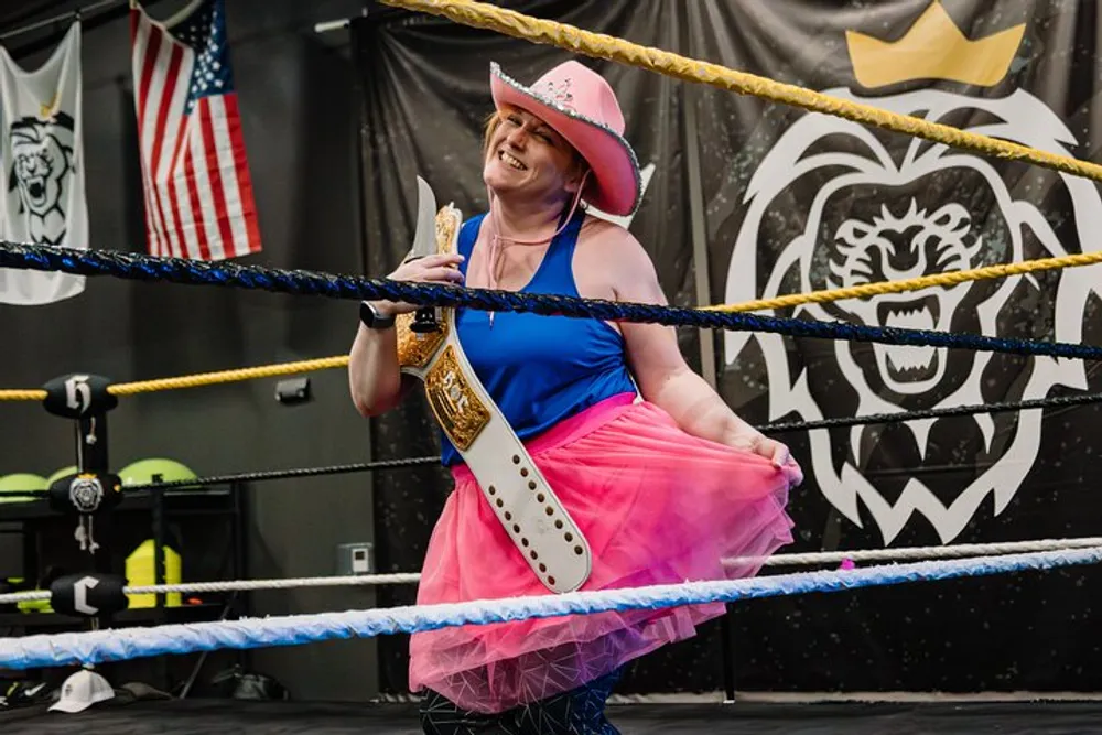 A smiling person in a vibrant blue top pink tutu and pink cowboy hat stands proudly in a wrestling ring with a championship belt over their shoulder