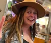 A smiling person wearing a hat makes a peace sign with one hand while holding a glass of beer in a lively bar setting