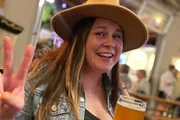 A smiling person wearing a hat makes a peace sign with one hand while holding a glass of beer in a lively bar setting.