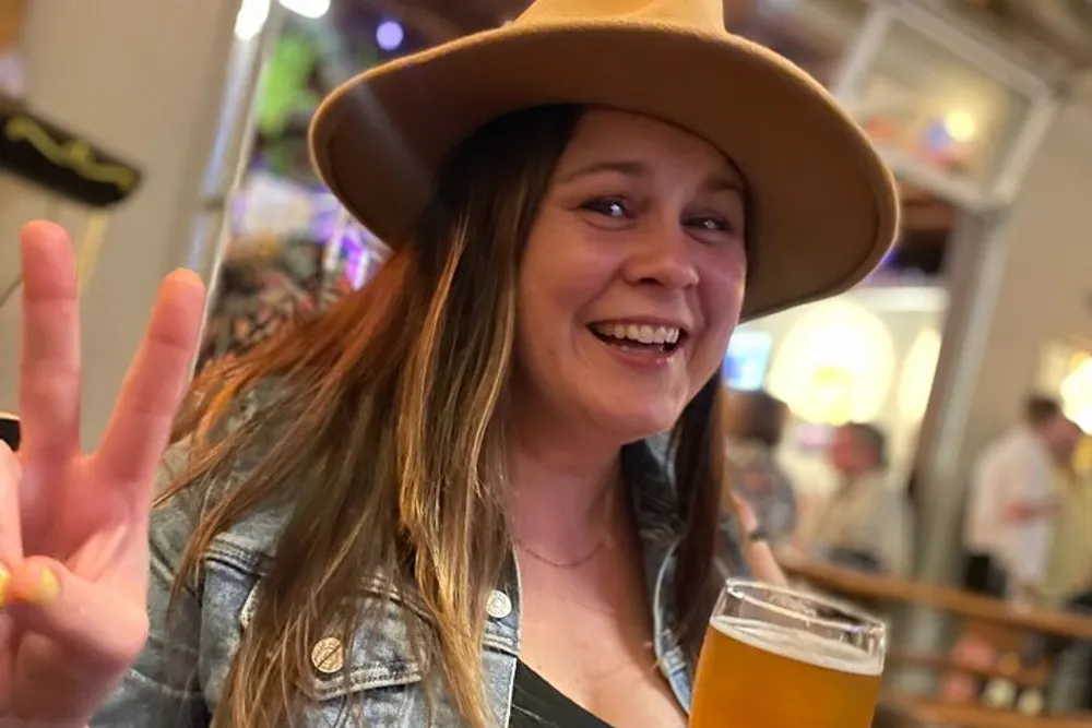 A smiling person wearing a hat makes a peace sign with one hand while holding a glass of beer in a lively bar setting