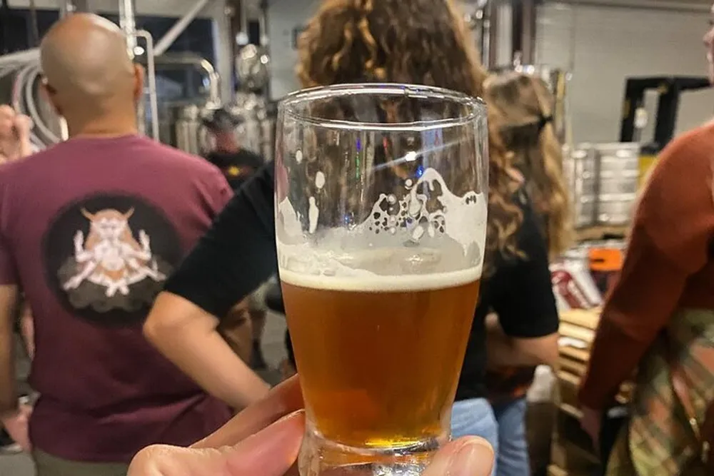 A person is holding a half-full glass of beer in focus with a group of people socializing in the blurry background possibly in a brewery