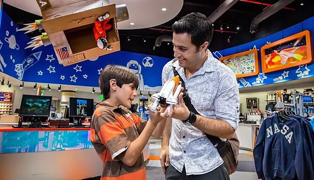 A man and a young boy are sharing a joyful moment as they look at a space shuttle model in a store with space-themed decor and NASA merchandise