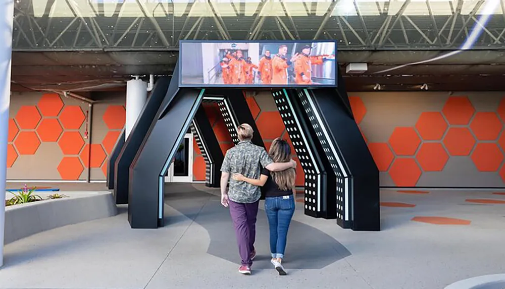 Two people are walking arm in arm looking at a screen displaying astronauts above a futuristic-looking structure with an orange hexagonal pattern on the wall behind them