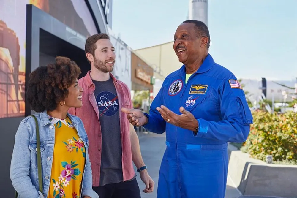 A person in a NASA astronaut flight suit is engaging in a lively conversation with two smiling individuals wearing casual clothes possibly sharing experiences or discussing space-related topics