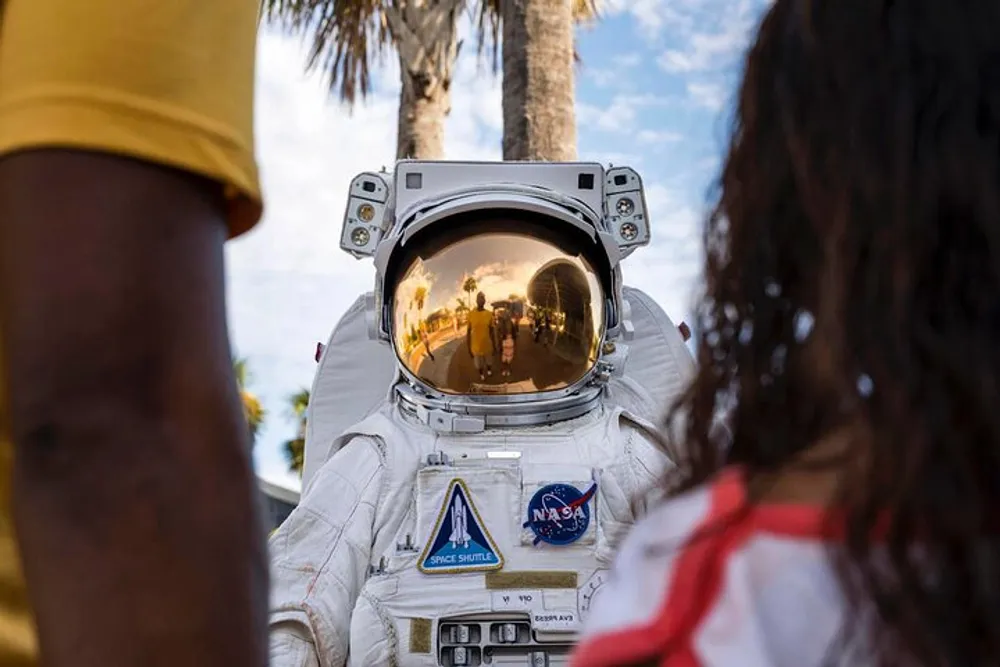 The image depicts a person in a NASA space suit engaging with two individuals amid a backdrop of tropical palm trees with the reflections in the helmet visor offering a glimpse of the surrounding environment