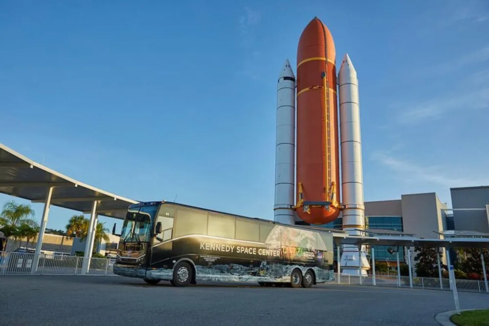 A tour bus adorned with Kennedy Space Center graphics is parked in front of a large display of a space shuttles external fuel tank and solid rocket boosters