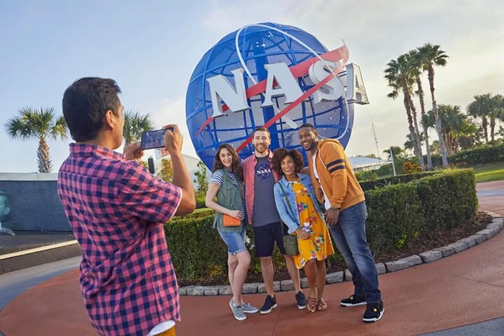 A person is taking a photo of three smiling friends posing in front of a large NASA emblem at a visitor center during the daytime