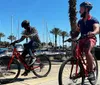 Two people are riding bicycles on a sunny day near a marina with palm trees and boats in the background