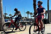 Two people are riding bicycles on a sunny day near a marina with palm trees and boats in the background.