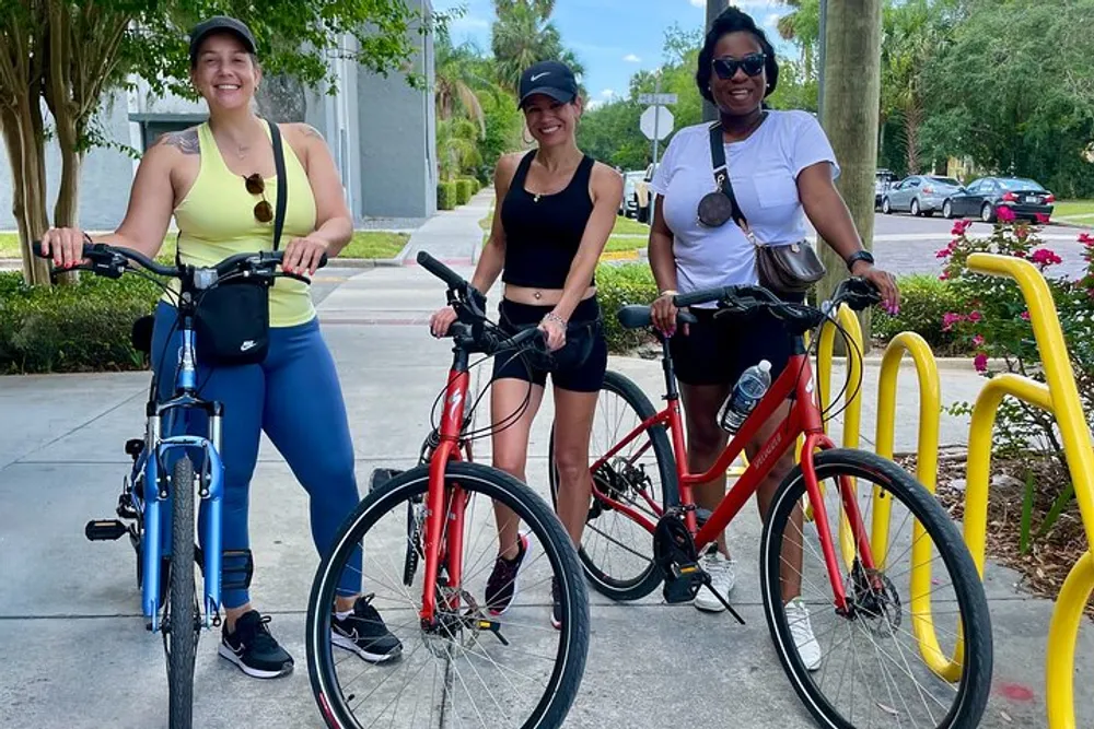 Three smiling women are standing with their bicycles ready for a ride in a sunny outdoor setting with greenery and yellow bike racks in the background