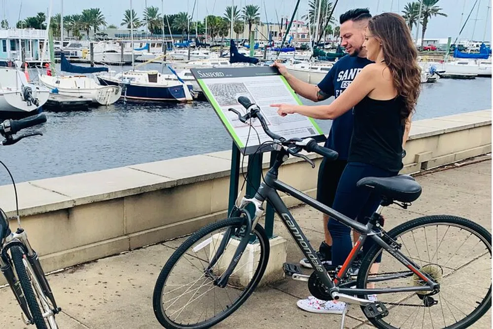 Two people are reading an information board beside a bicycle with a marina full of boats in the background
