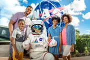 A family is posing with a person in an astronaut suit in front of a large NASA sign under a sunny sky.