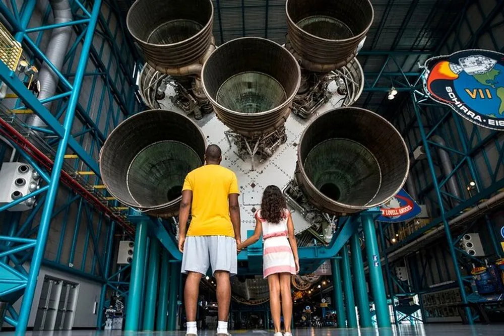 Two people are holding hands and looking up at the immense engines of a space rocket possibly on display in a museum or visitor center