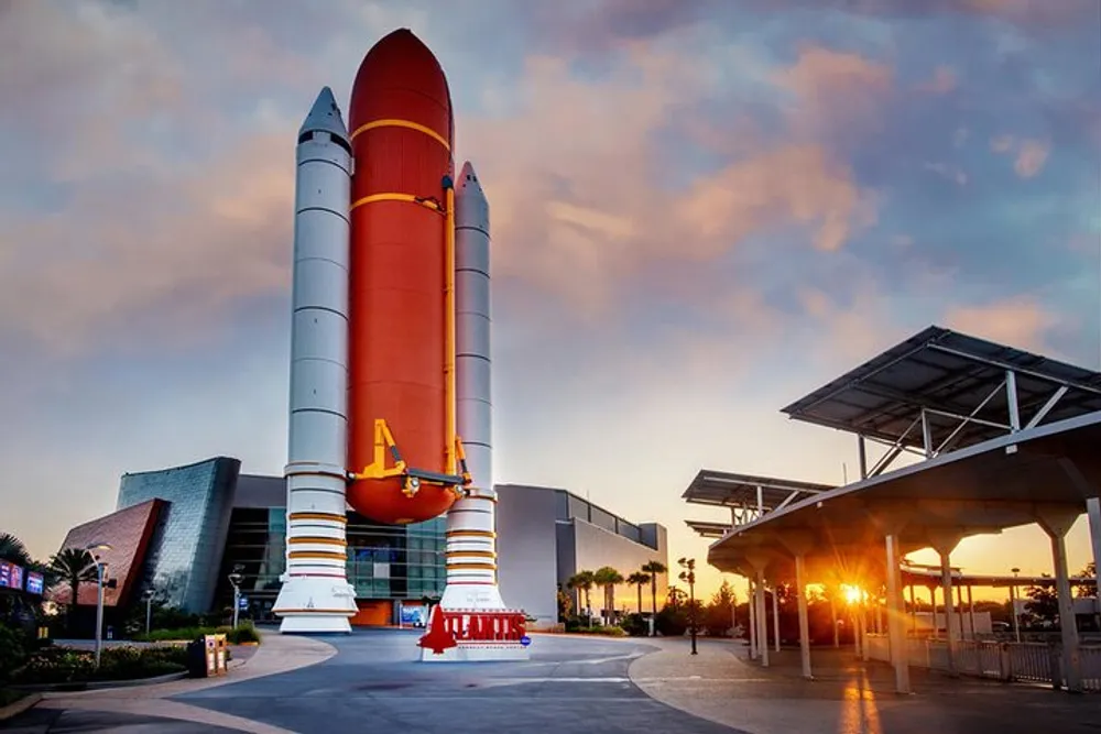 The image shows a large Space Shuttle exhibit with its external fuel tank and solid rocket boosters on display at a museum during sunset