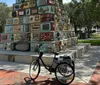 A bicycle with a basket is parked in front of a unique multi-colored mosaic tower in a sunny outdoor setting