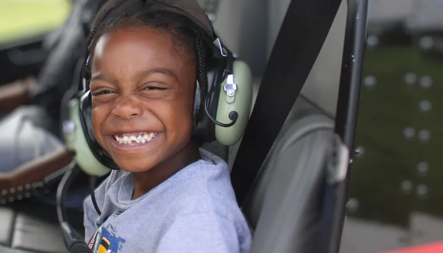 A joyful child is wearing aviation headphones and smiling widely inside an aircraft.
