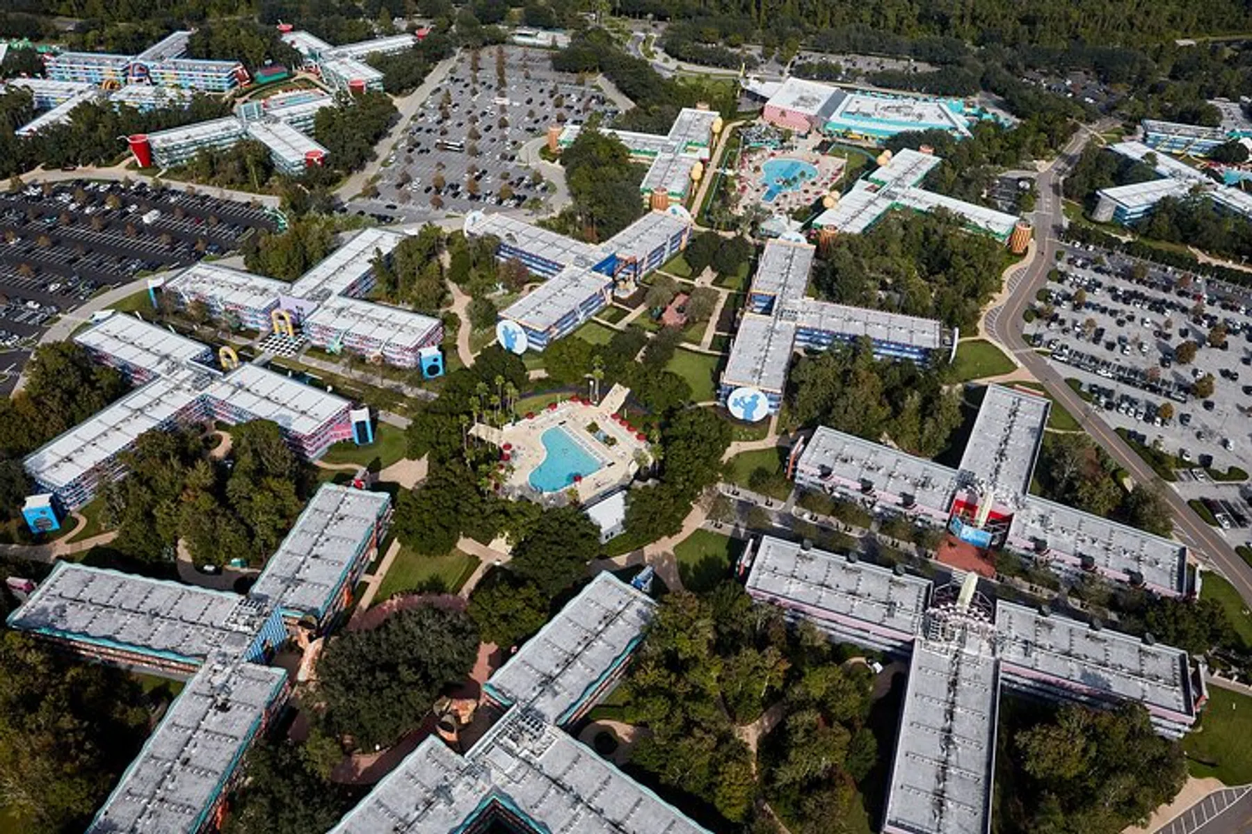 The image is an aerial view of a large resort complex featuring multiple buildings with colorful rooftops, swimming pools, and spacious parking areas amidst greenery.