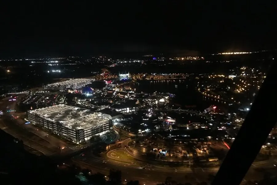 An aerial night view of a vibrant cityscape with illuminated streets, buildings, and possibly a fairgrounds or amusement park near a body of water.